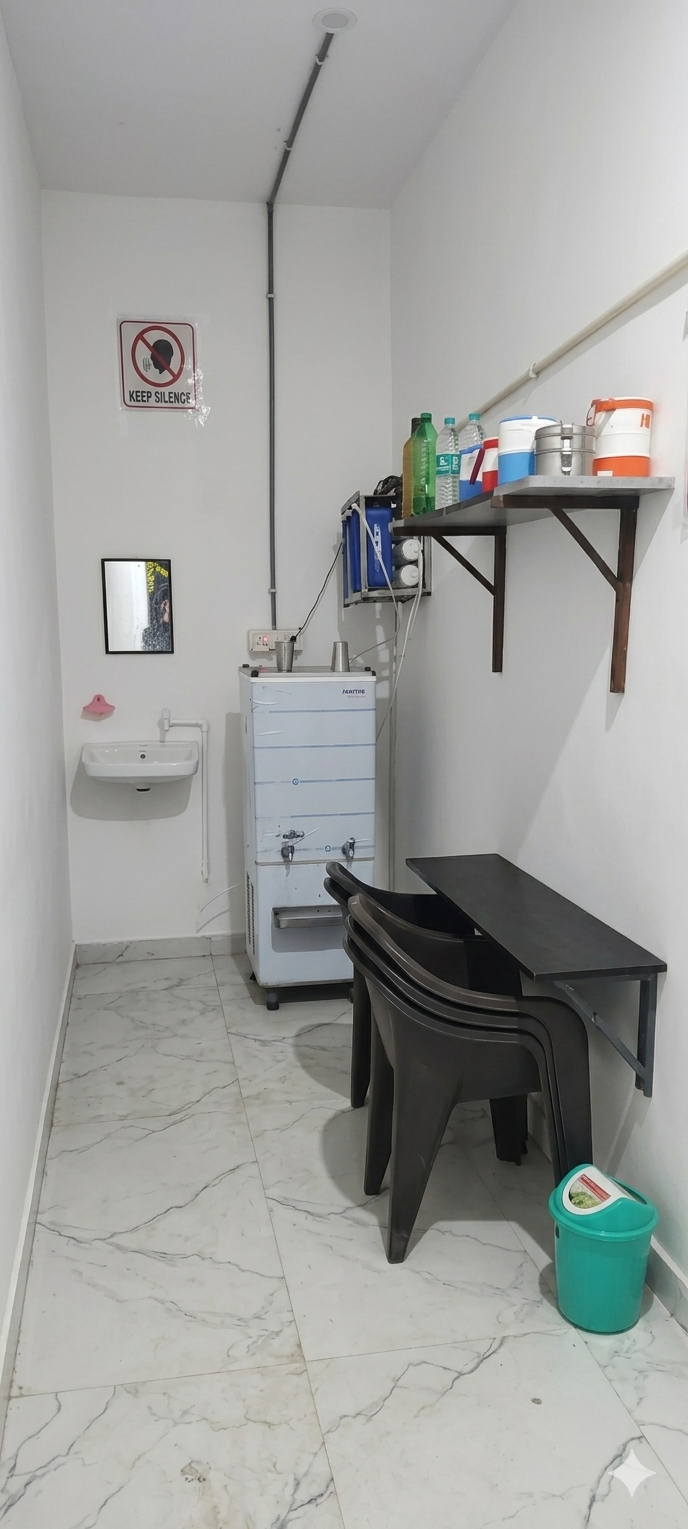 Clean refreshment and utility zone outside the library's main study room. Features an RO water cooler, washbasin, wall-mounted dining table, and seating area for student tiffin breaks.